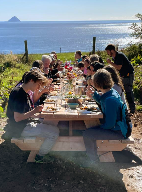 Grupo de gente de diversas edades sentada alrededor de la mesa en zona verde enfrente del mar